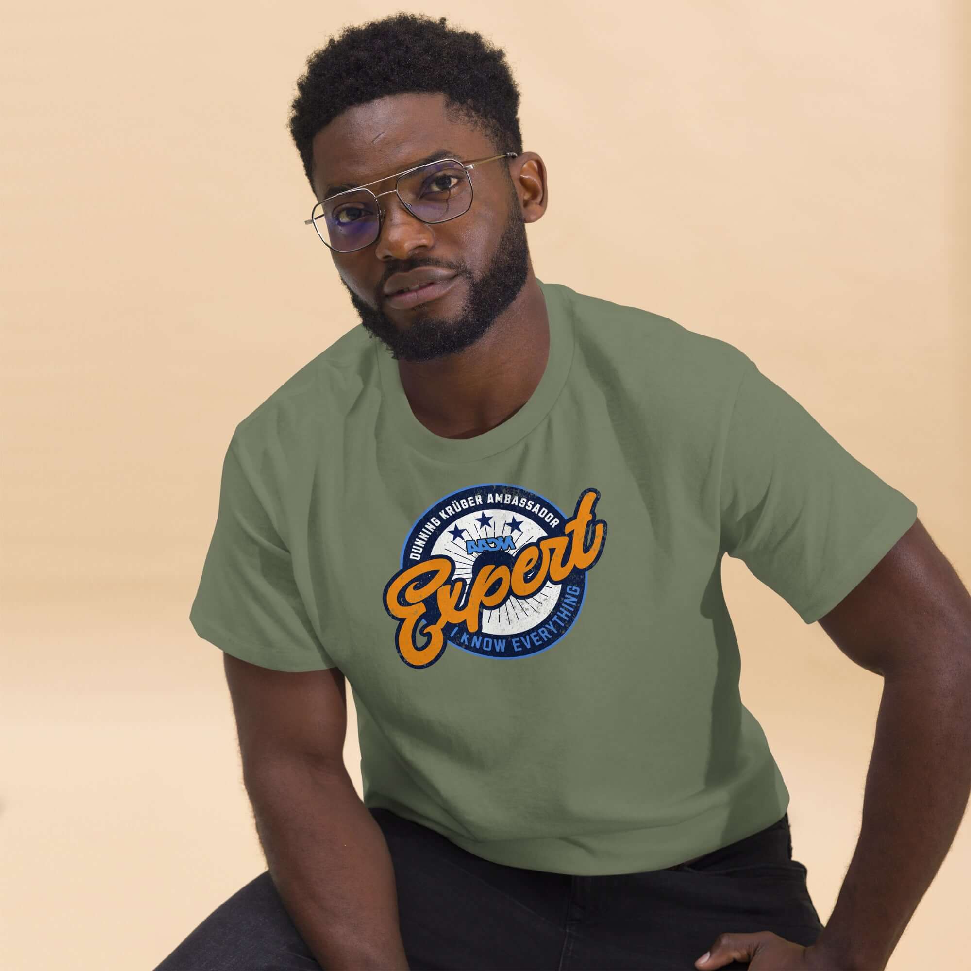 Model wearing olive-green Dunning-Kruger Expert T-shirt with a circular emblem, promoting humorous take on psychology and self-perception.