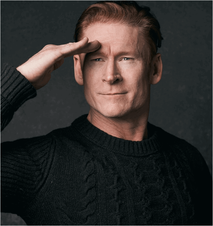 Actor Zack Ward saluting, wearing a black sweater against a dark background.
