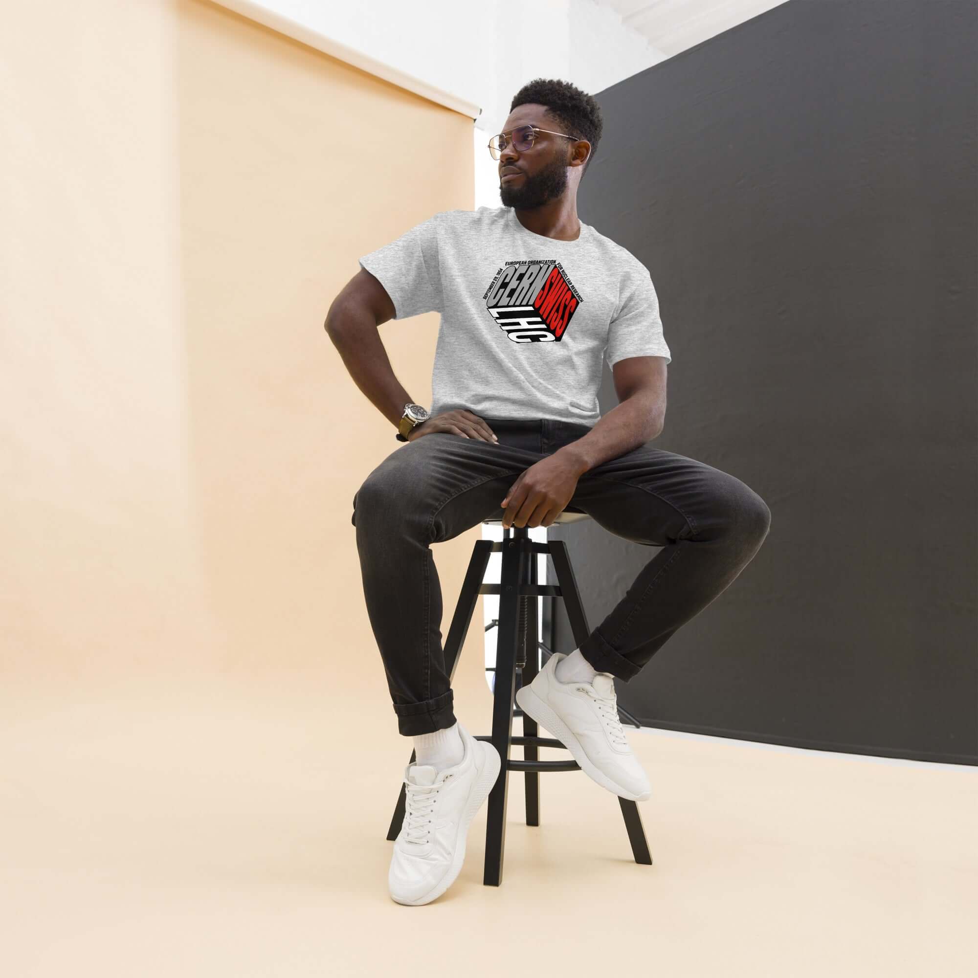 Man wearing CERN-themed physics cube t-shirt, styled with dark jeans and white sneakers, sitting on a stool in a modern studio.