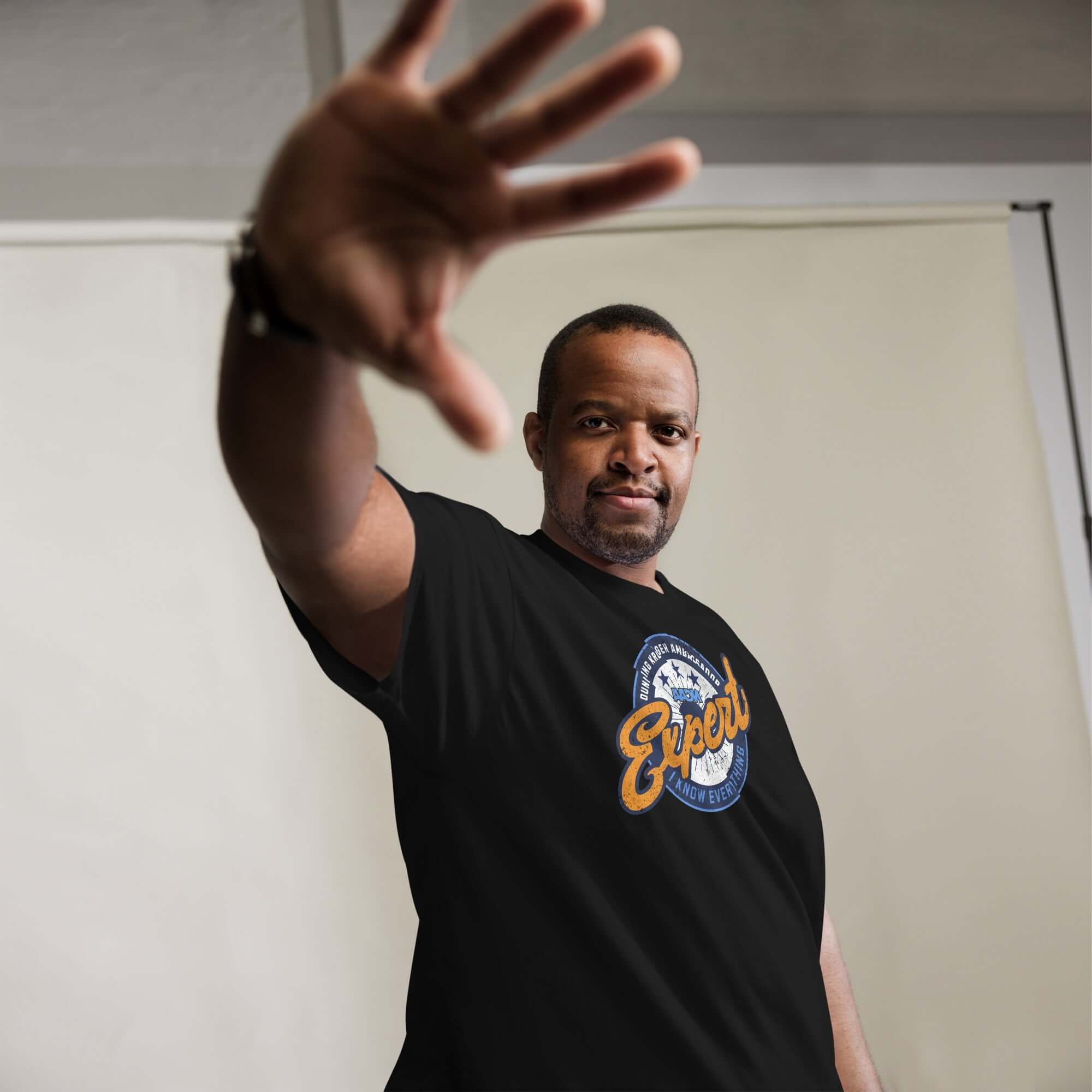 Man wearing black Dunning-Kruger Expert T-Shirt with circular emblem, extending hand towards camera, posing confidently indoors.