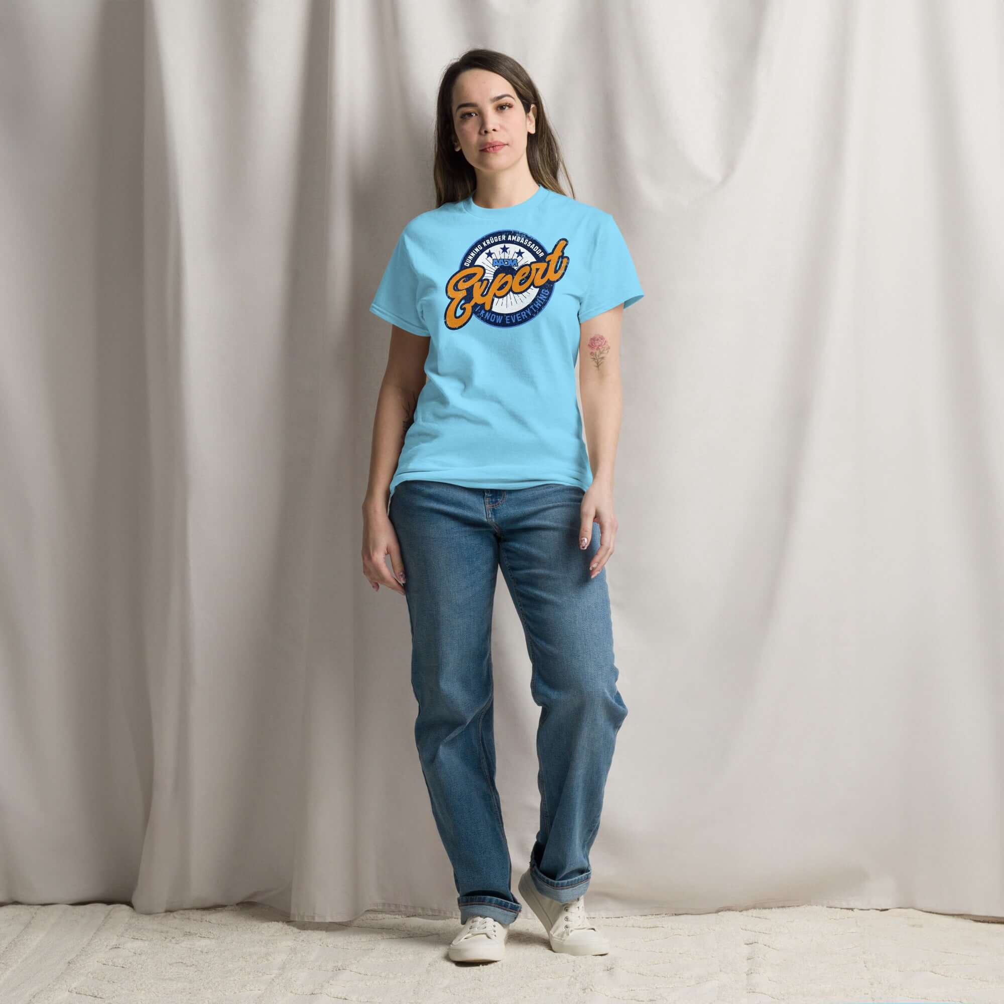 Person modeling light blue Dunning-Kruger Expert T-Shirt with sports logo design, standing against a neutral backdrop.