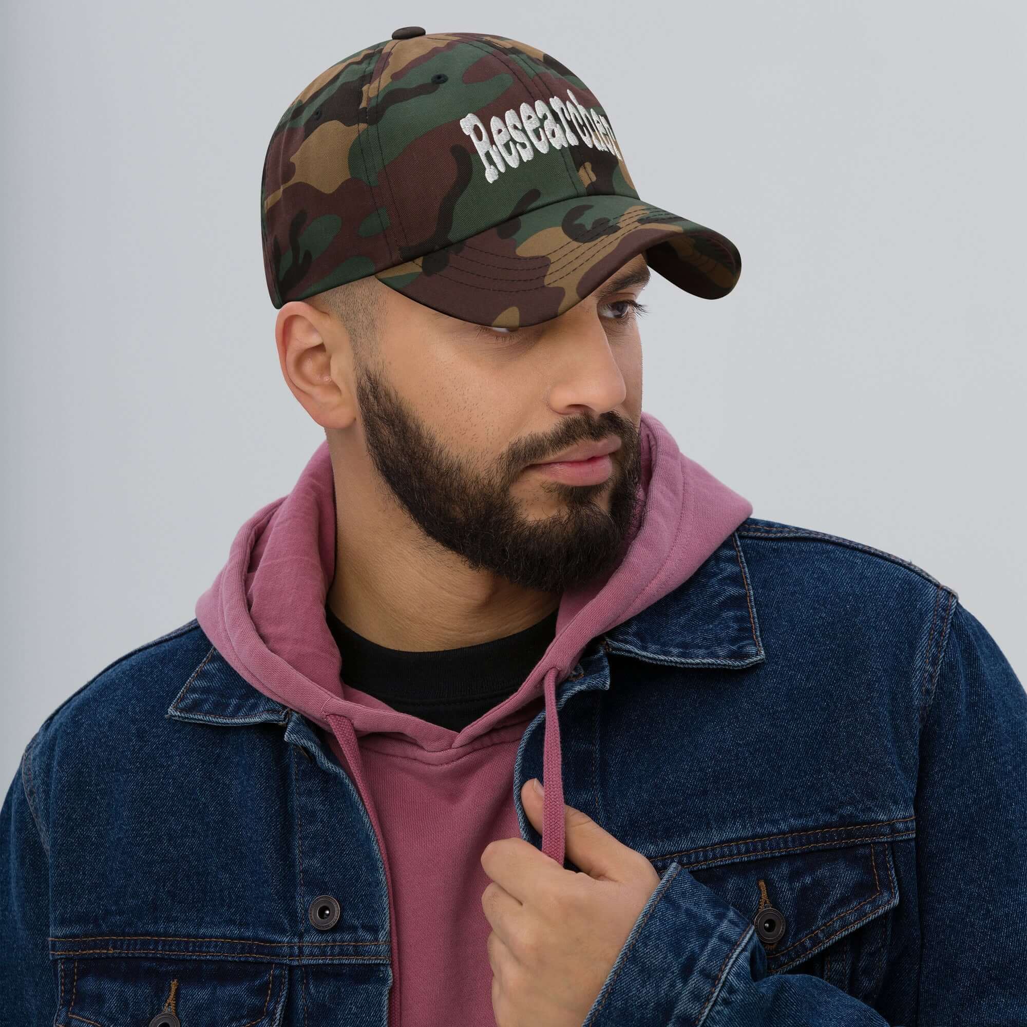 Camouflage "Researcher" baseball cap on person wearing a pink hoodie and denim jacket, symbolizing curiosity and professionalism.