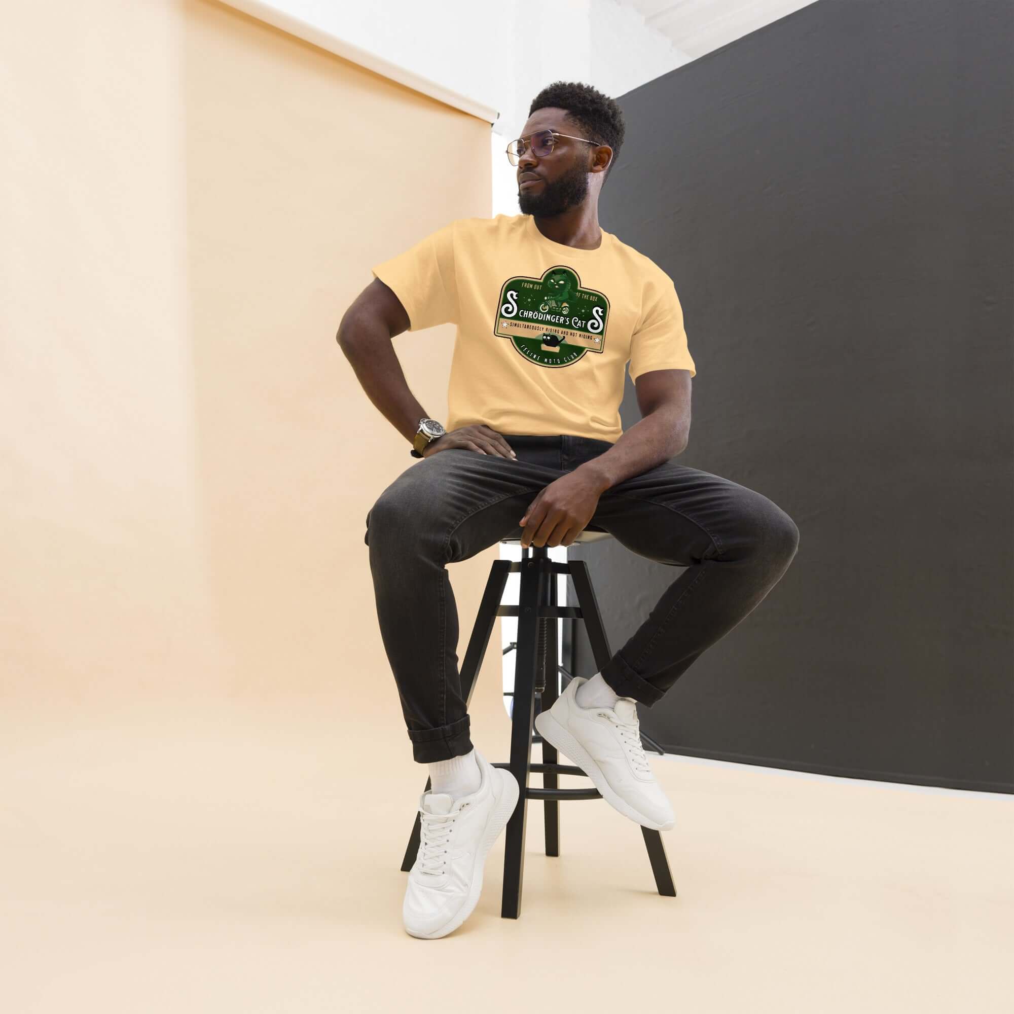Man in yellow Schrödinger's Cats Motorcycle Club T-shirt sitting on stool, wearing black jeans and white sneakers, studio background.